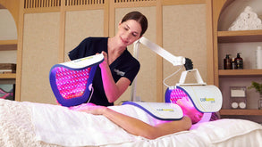 Esthetician placing a panel onto a clients hands while they lie on a massage table with additional panels over their neck/chest and face from the Celluma ELITE device