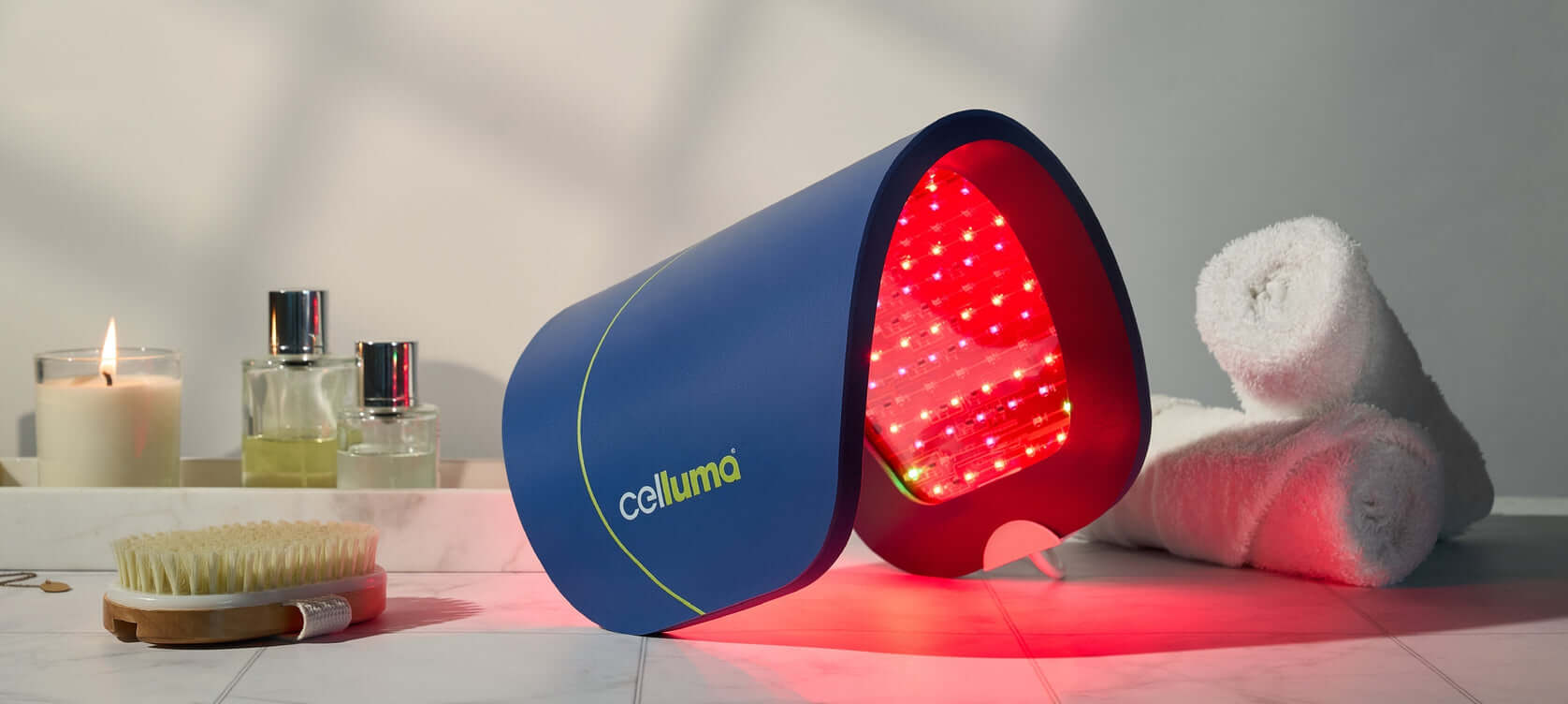 A Celluma panel in a curved shape sitting on a table with two small, rolled towels, a body brush, a lit candle, and two bottles of body oil.