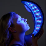 Person holding a crescent-shaped Celluma NOVA blue and red light therapy LED panel in front of their face against a dark background.